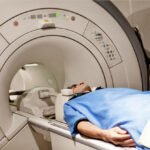 Young man in blue protective hospital clothes having medical examination via MRI scan.The focus is on model.The photo is taken with a full frame DSLR camera in horizontal composition.The man is in his thirties and has brown hair located on the right of frame.